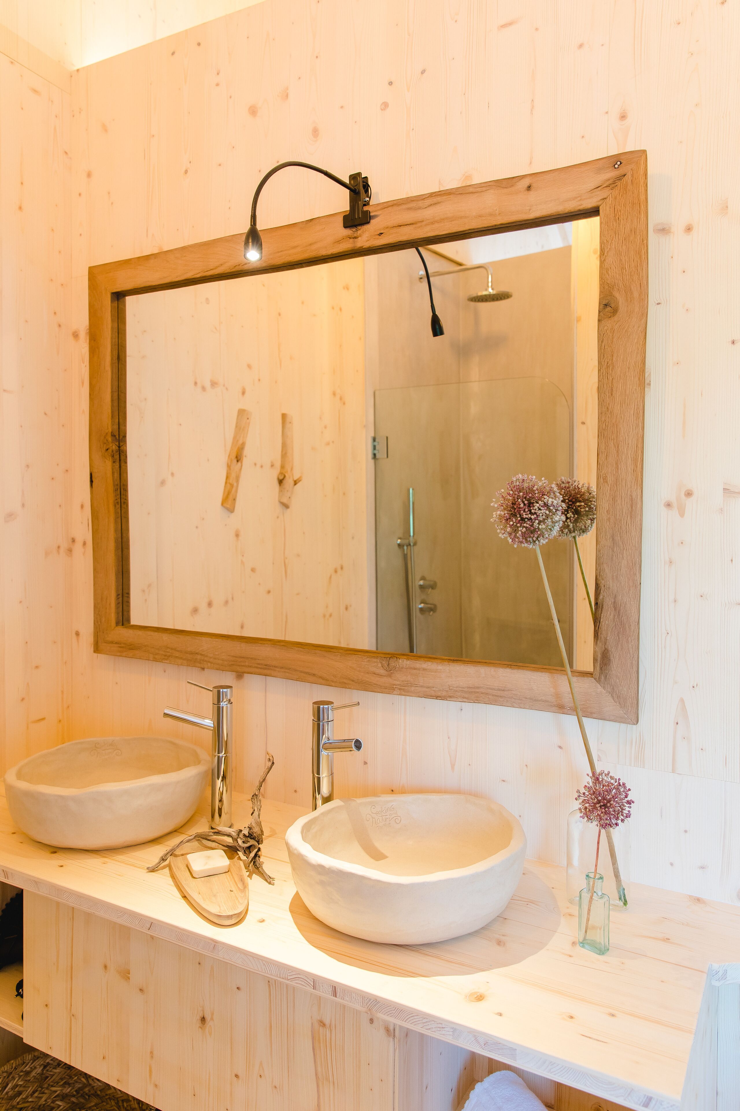 Un baño de madera con dos lavamanos, un espejo y un pequeño jarrón con flores.