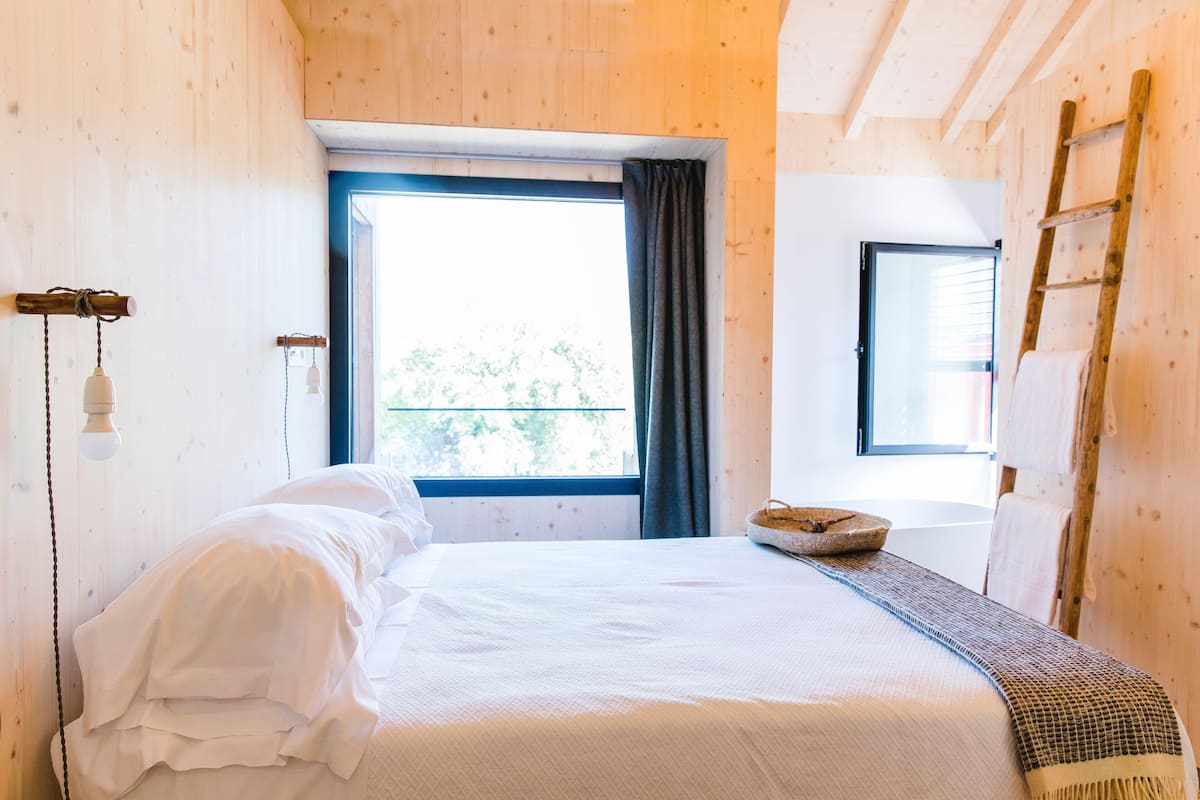 A wooden-paneled room with a bed, a window with curtains, a ladder, and a hanging light fixture.
