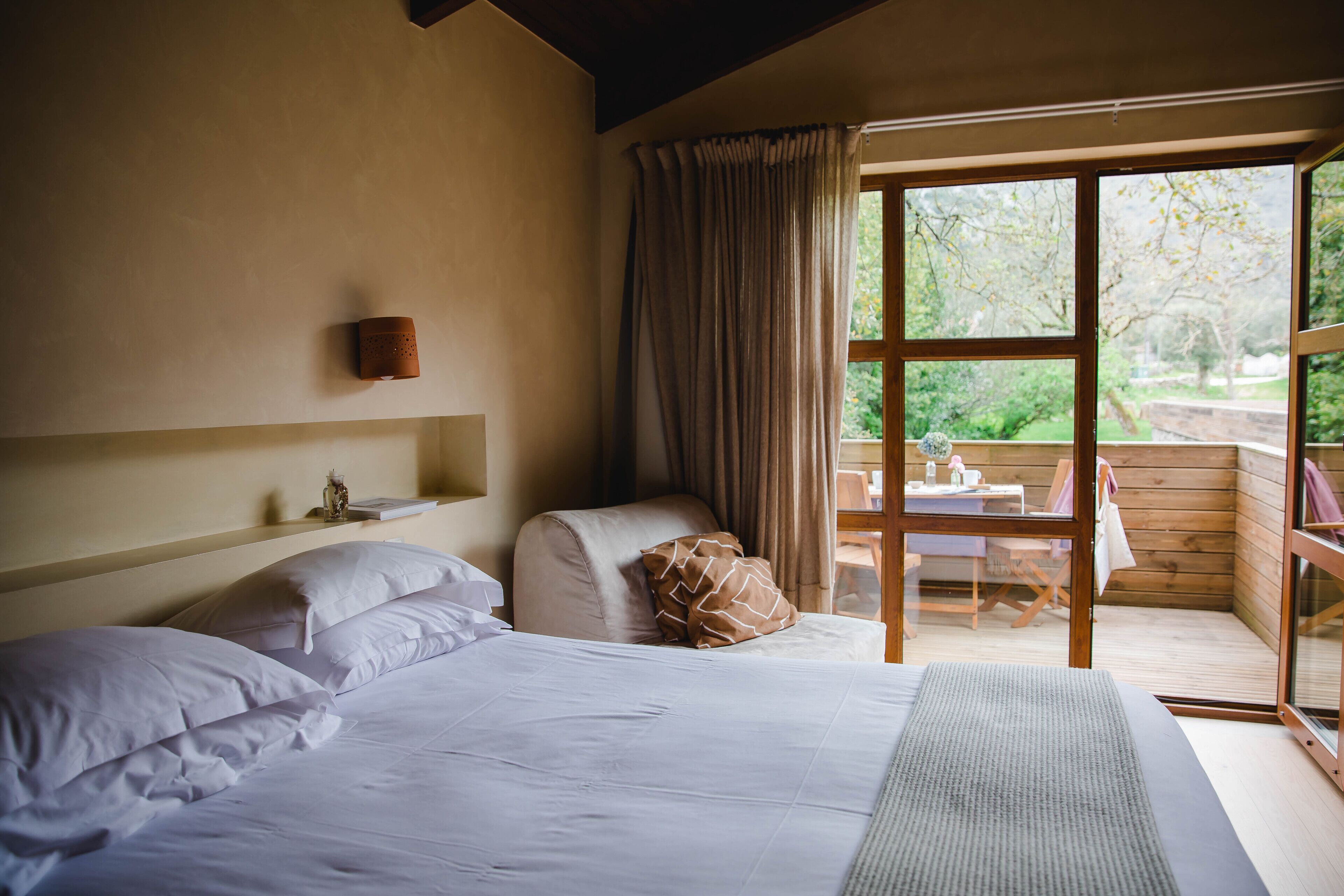 A bedroom with a large bed, a wooden headboard, a small table with a chair, and a view of the outdoors through a glass door.