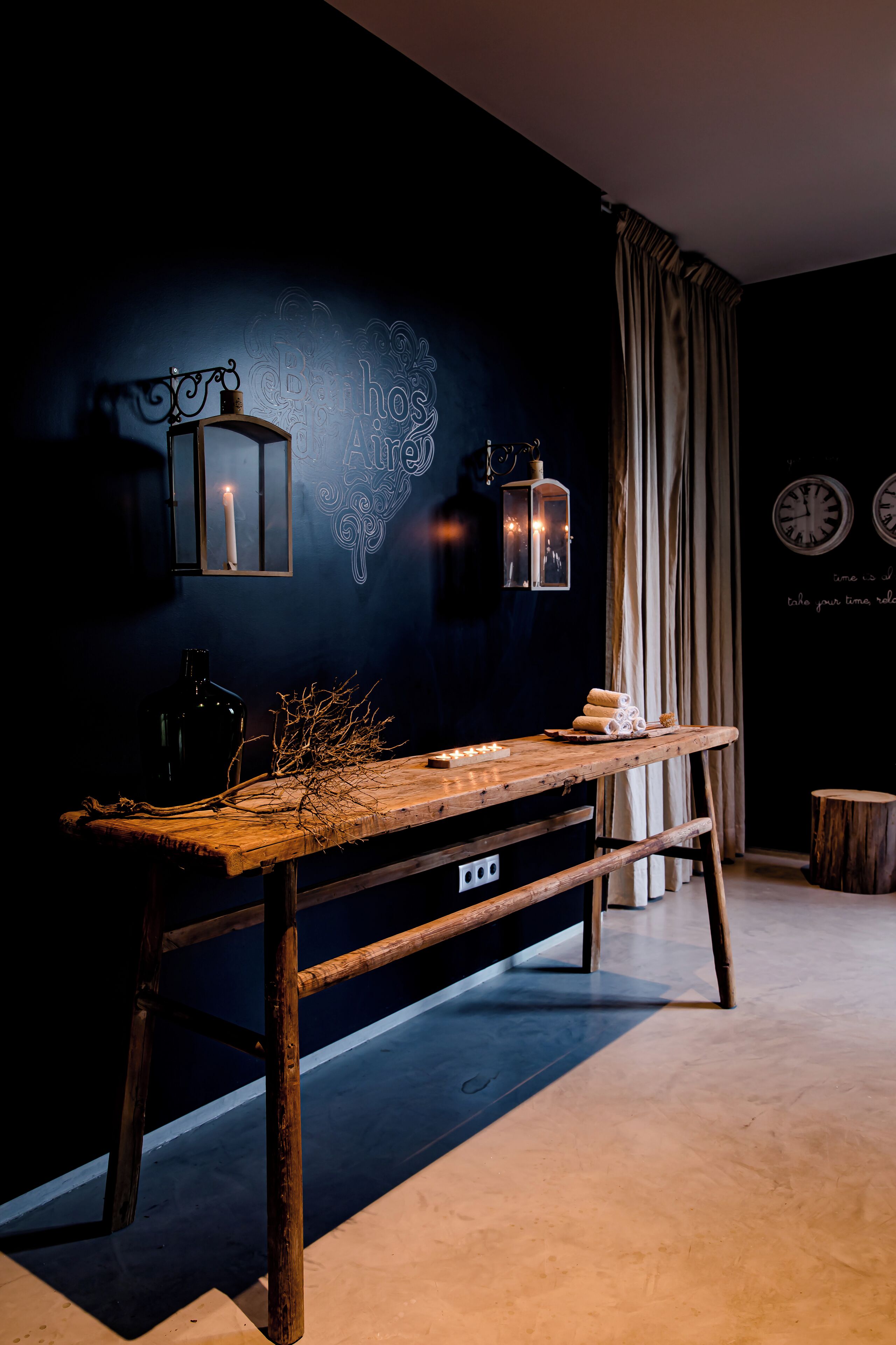 Een kamer in donkere tinten met een houten tafel, kaarsen en lantaarns aan de muur.