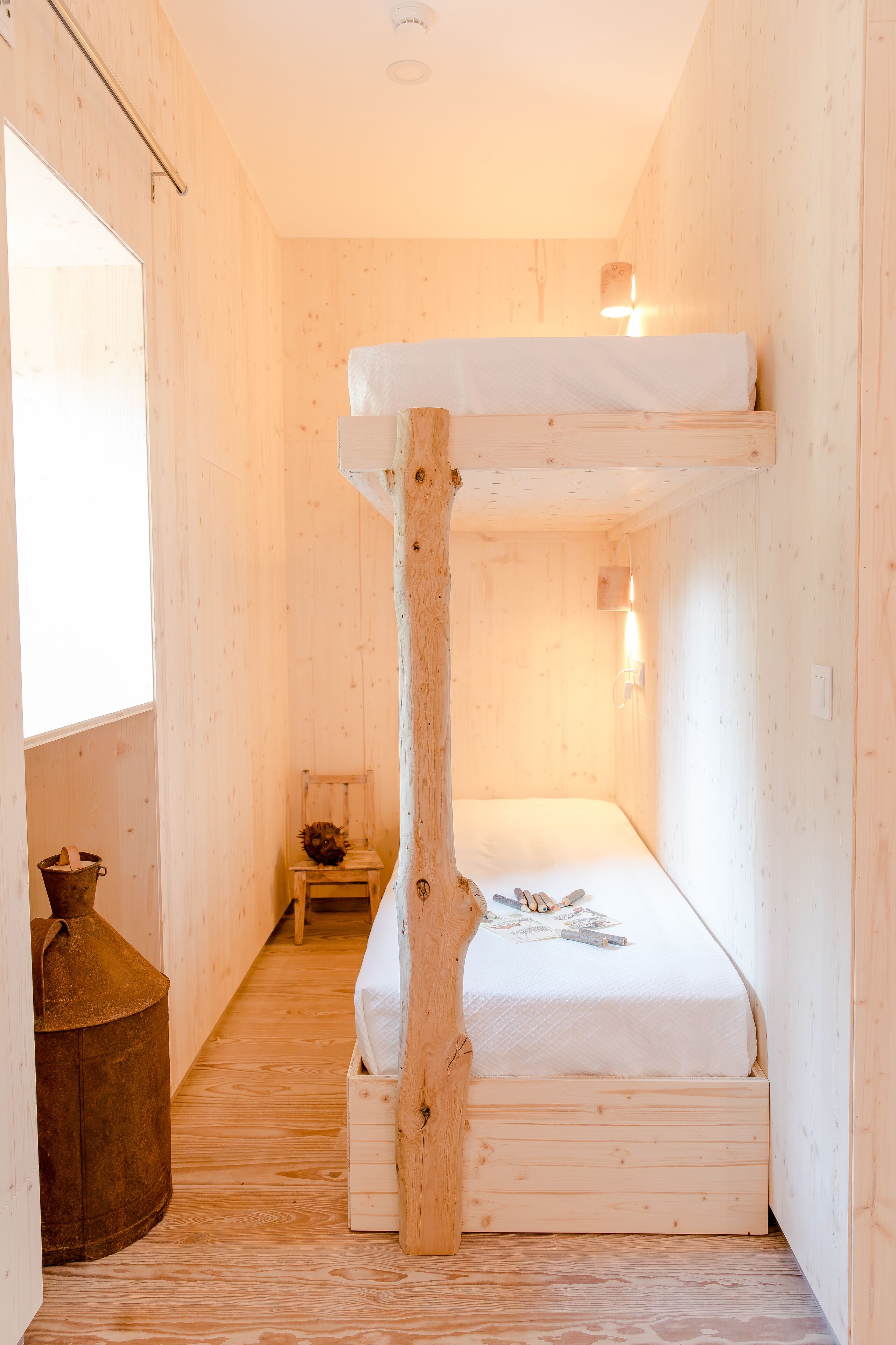 A wooden bunk bed with a tree trunk design, a small table, and a window in a cabin-style room.
