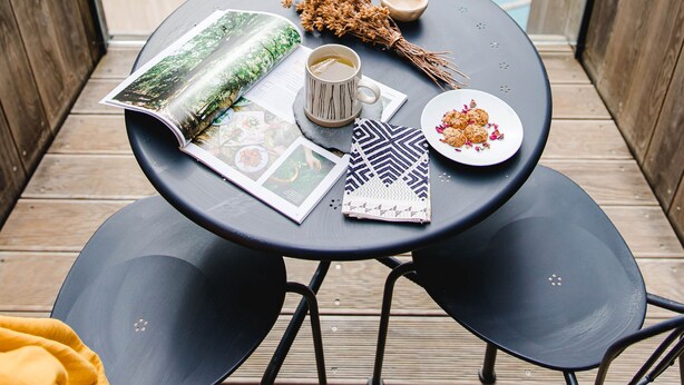 Une table ronde sur laquelle se trouvent un magazine, une tasse, une assiette avec de la nourriture et un objet décoratif, le tout placé sur un balcon donnant sur une piscine et un espace verdoyant.