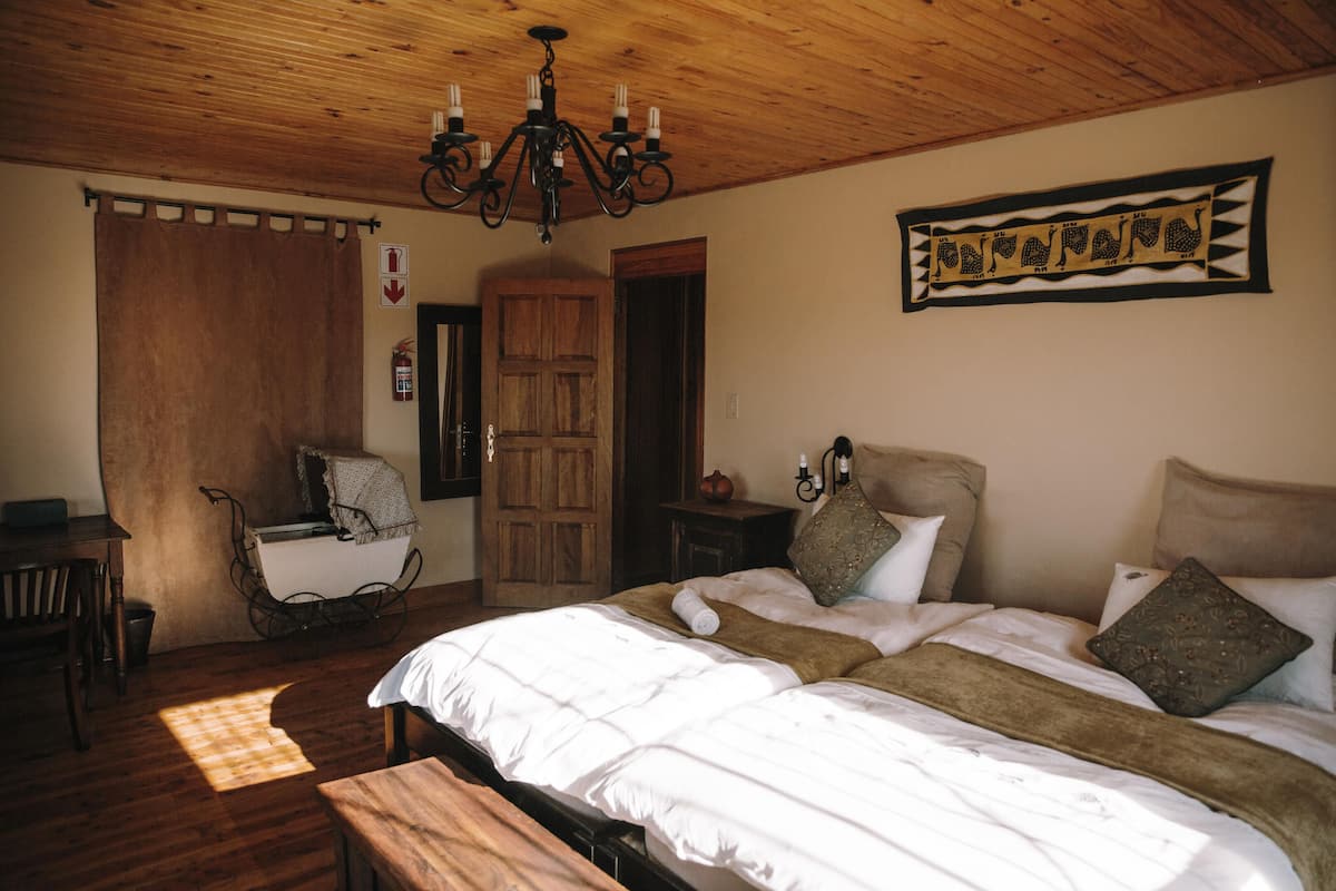 A bedroom with a wooden ceiling, a bed with white bedding, a wooden bench, a rocking chair, and a chandelier.