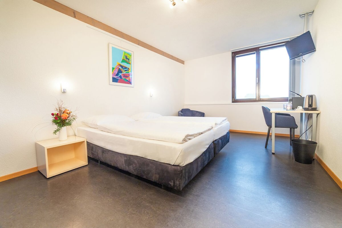 A modern hotel room with a bed, a desk, a chair, a small table, a trash bin, and a window with a view.