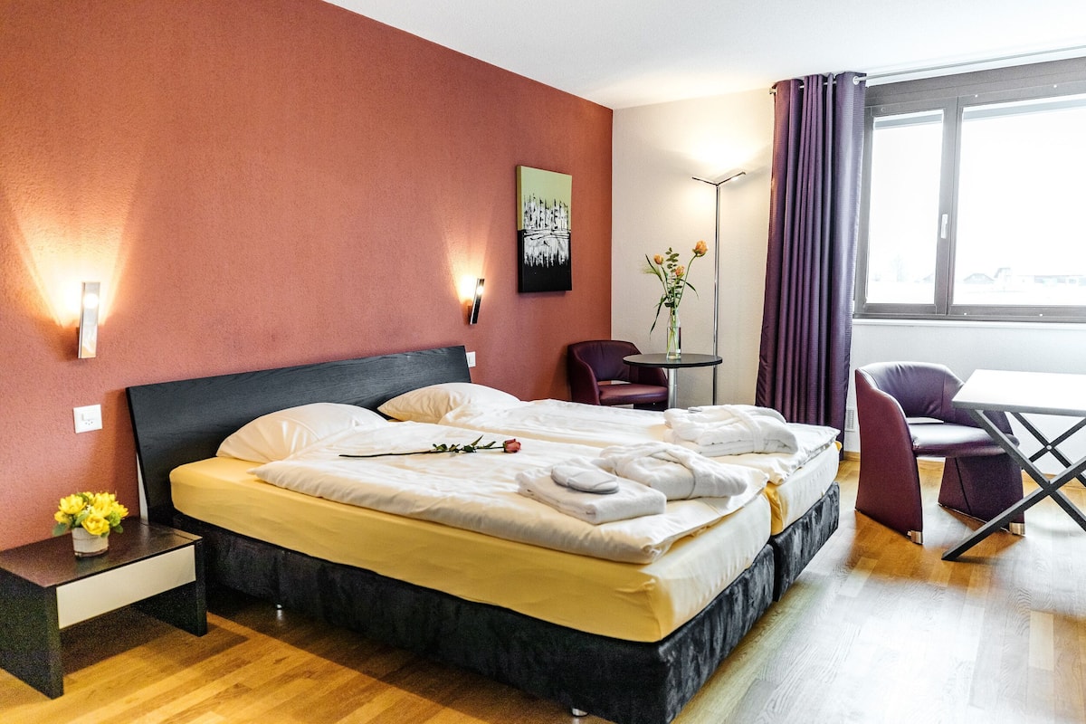 A hotel room with a bed, bedside table, chair, and a small table with a vase of flowers.