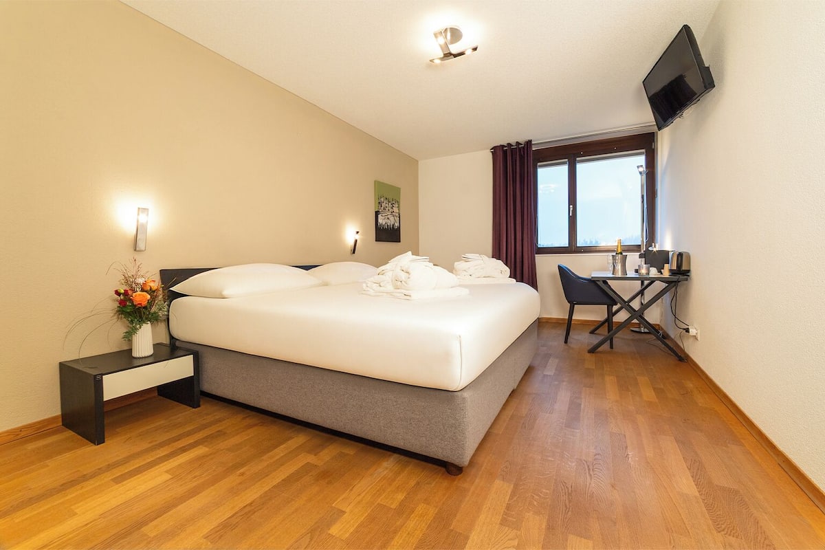 A hotel room with a bed, bedside table, chair, desk, TV, and a window with curtains.