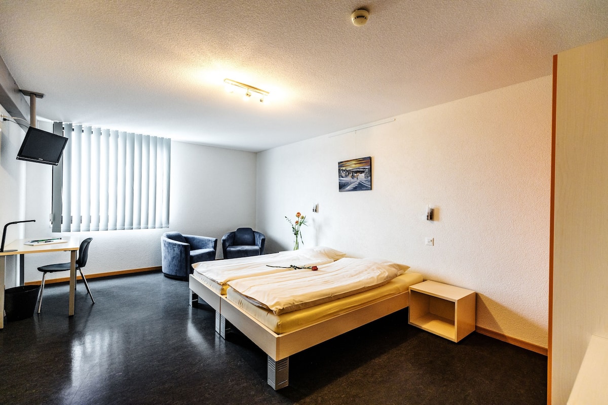A hotel room with a bed, desk, chair, TV, and a window with blinds.