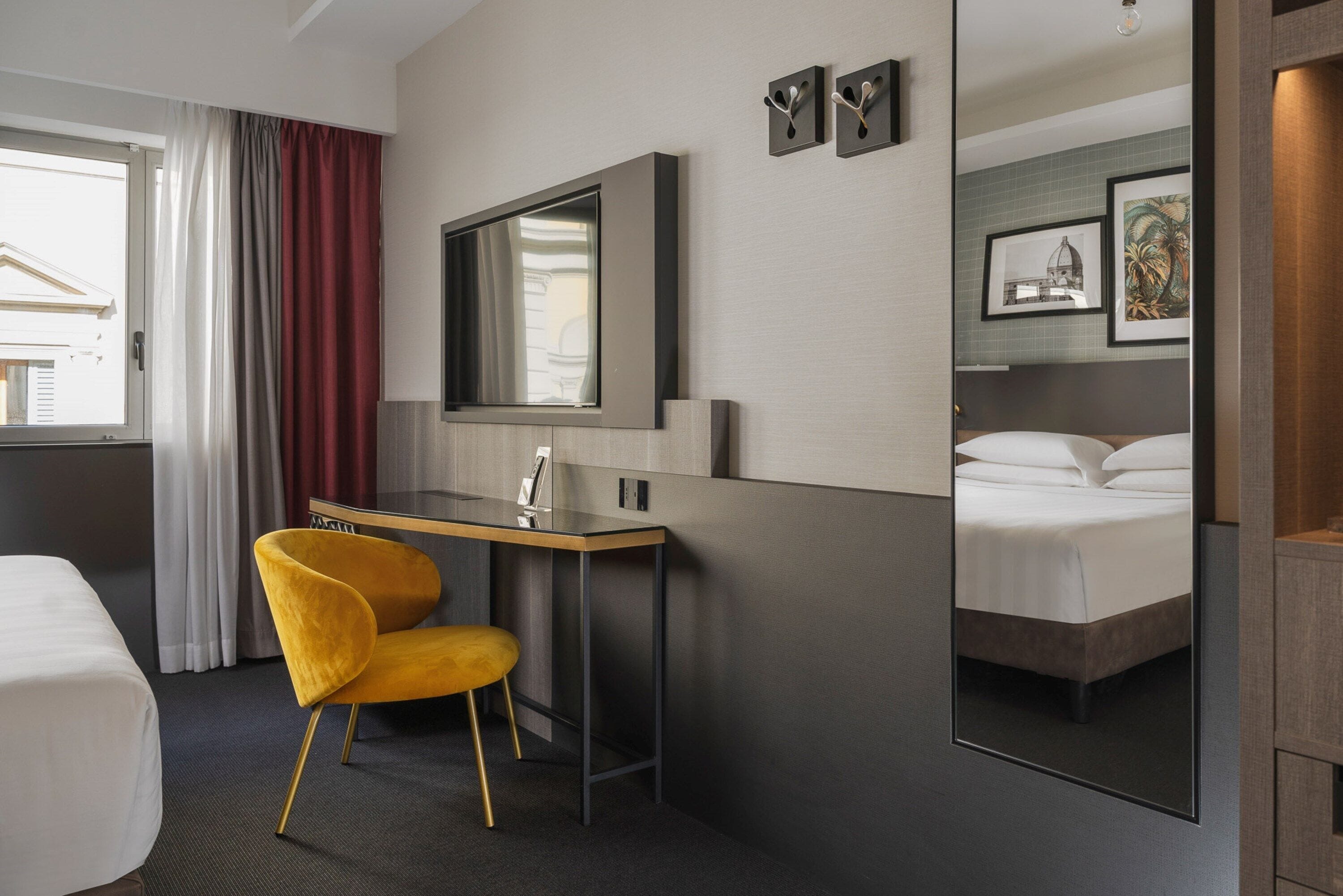 Una camera d'albergo con un letto, una scrivania, una sedia, uno specchio e una finestra con tende.