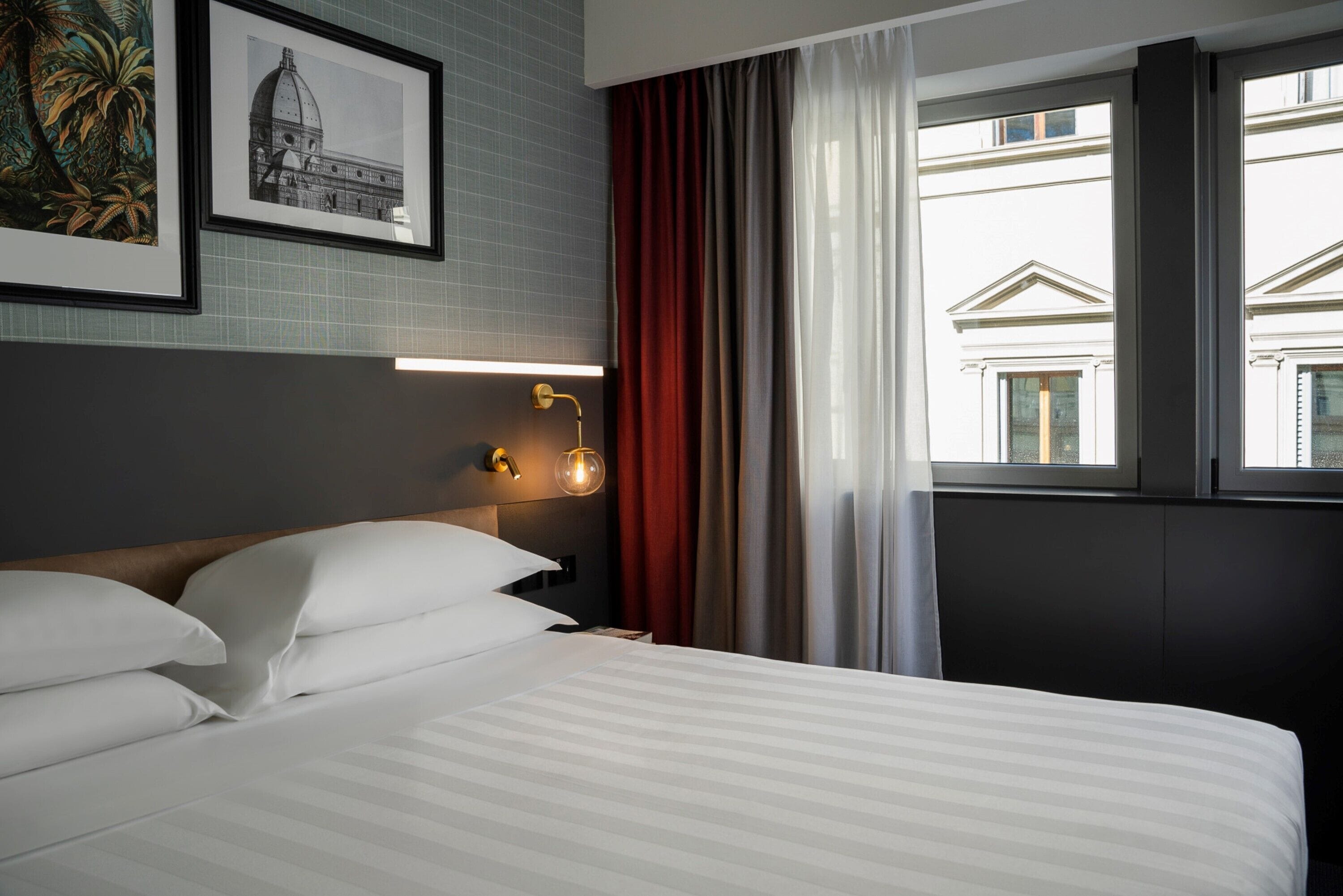 Una habitación de hotel con una cama bien hecha, una lámpara de pared y un ventanal con cortinas.