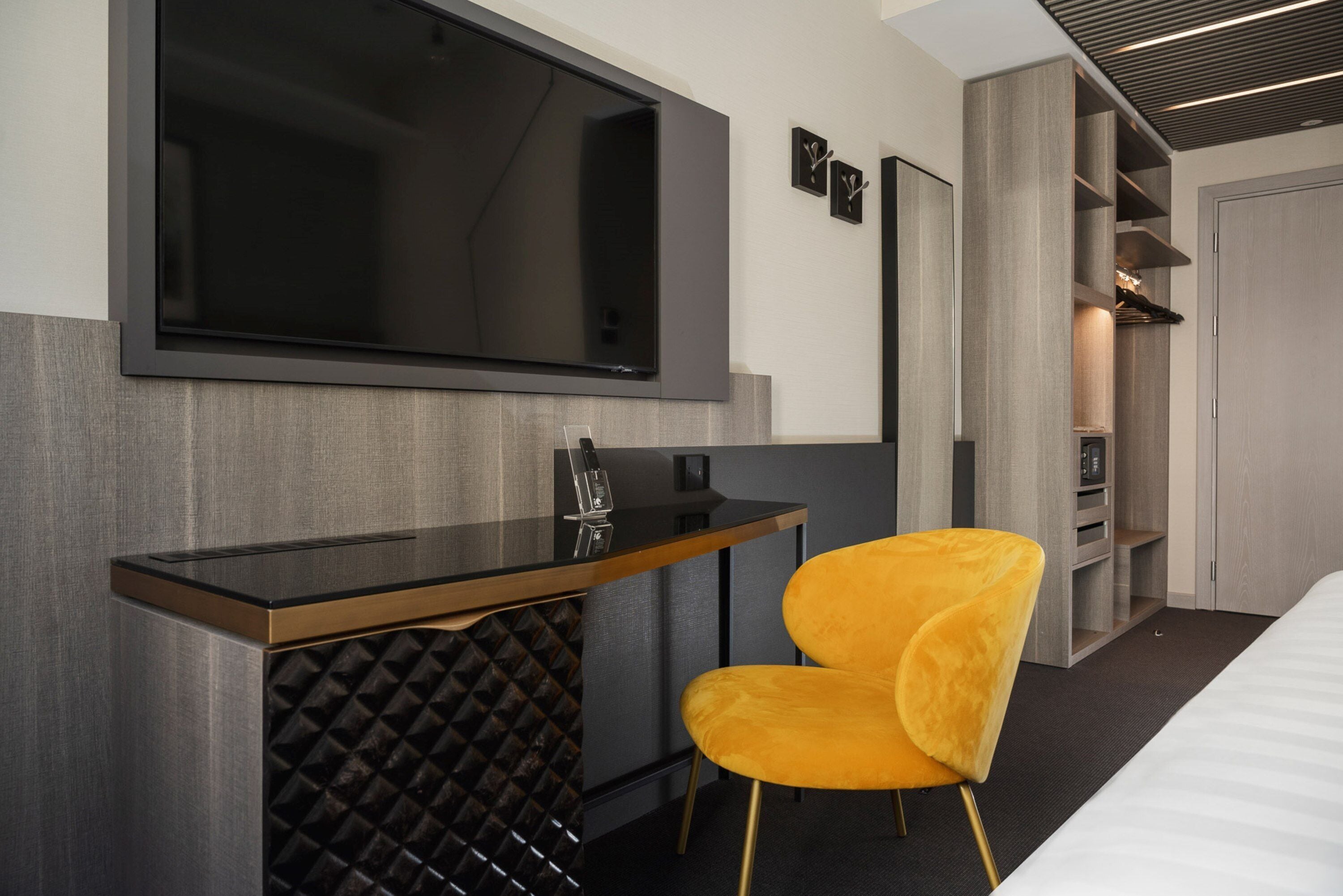 A modern hotel room with a large flat-screen TV mounted on the wall, a wooden desk with a chair, and a wardrobe with built-in shelving.
