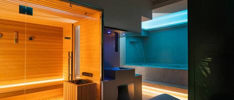 Een moderne sauna met houten wanden en een glazen omheining, een apart ontspanningsgedeelte met een massagetafel, en een waterpartij met blauwe verlichting.