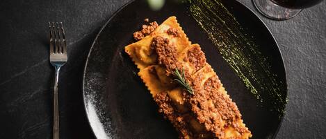 A plate of ravioli with a meat topping, a fork, and a glass of red wine on a dark surface.