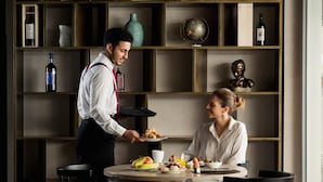 Un comedor con una mesa dispuesta para dos personas, un camarero sirviendo comida y una estantería al fondo.