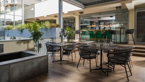 Un espacio de comedor al aire libre con diseño moderno, que incluye una mesa larga con sillas, un macetero con plantas y vistas al interior del edificio.