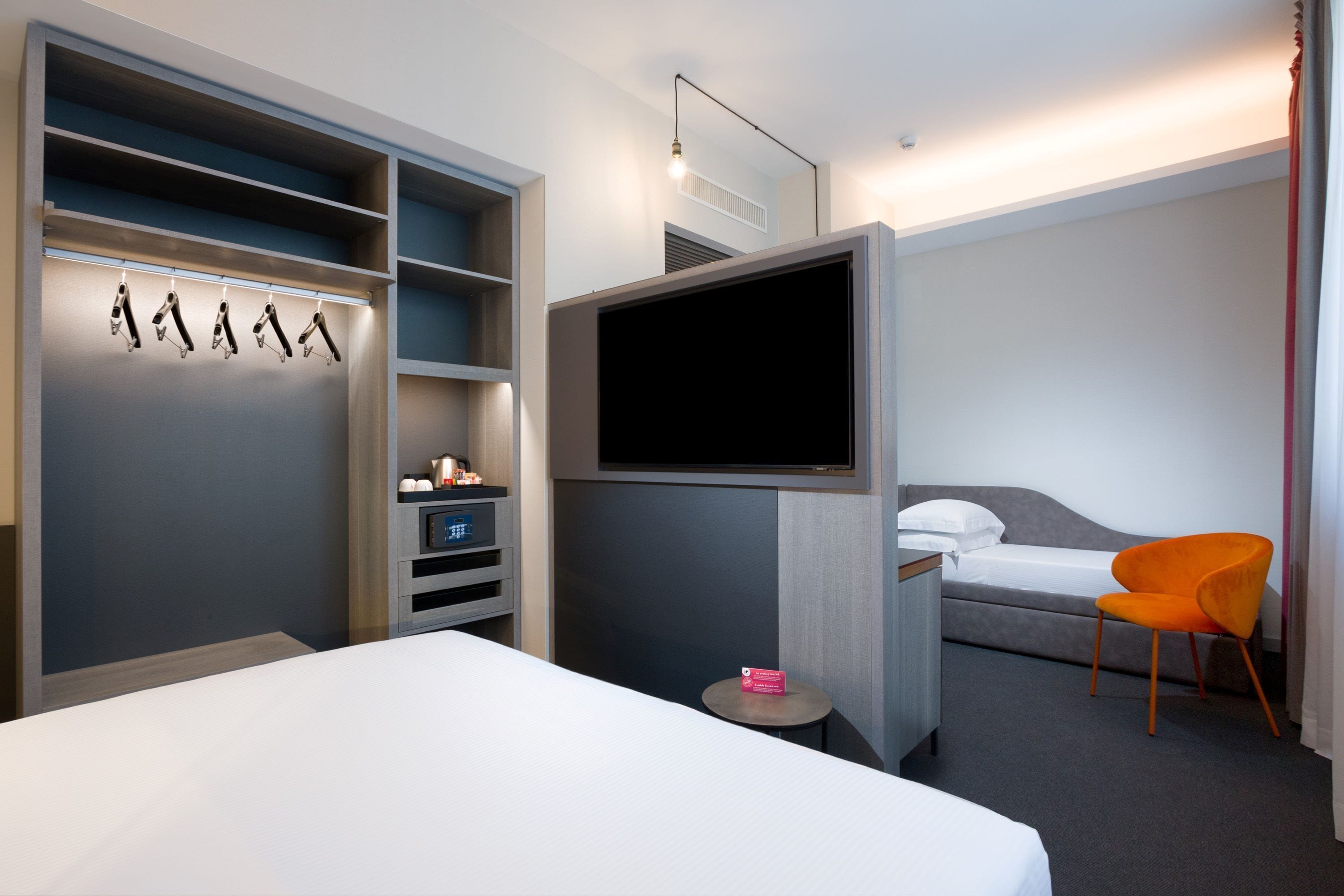 A modern hotel room with a bed, a TV, a desk, a chair, and a wardrobe with hanging shoes.
