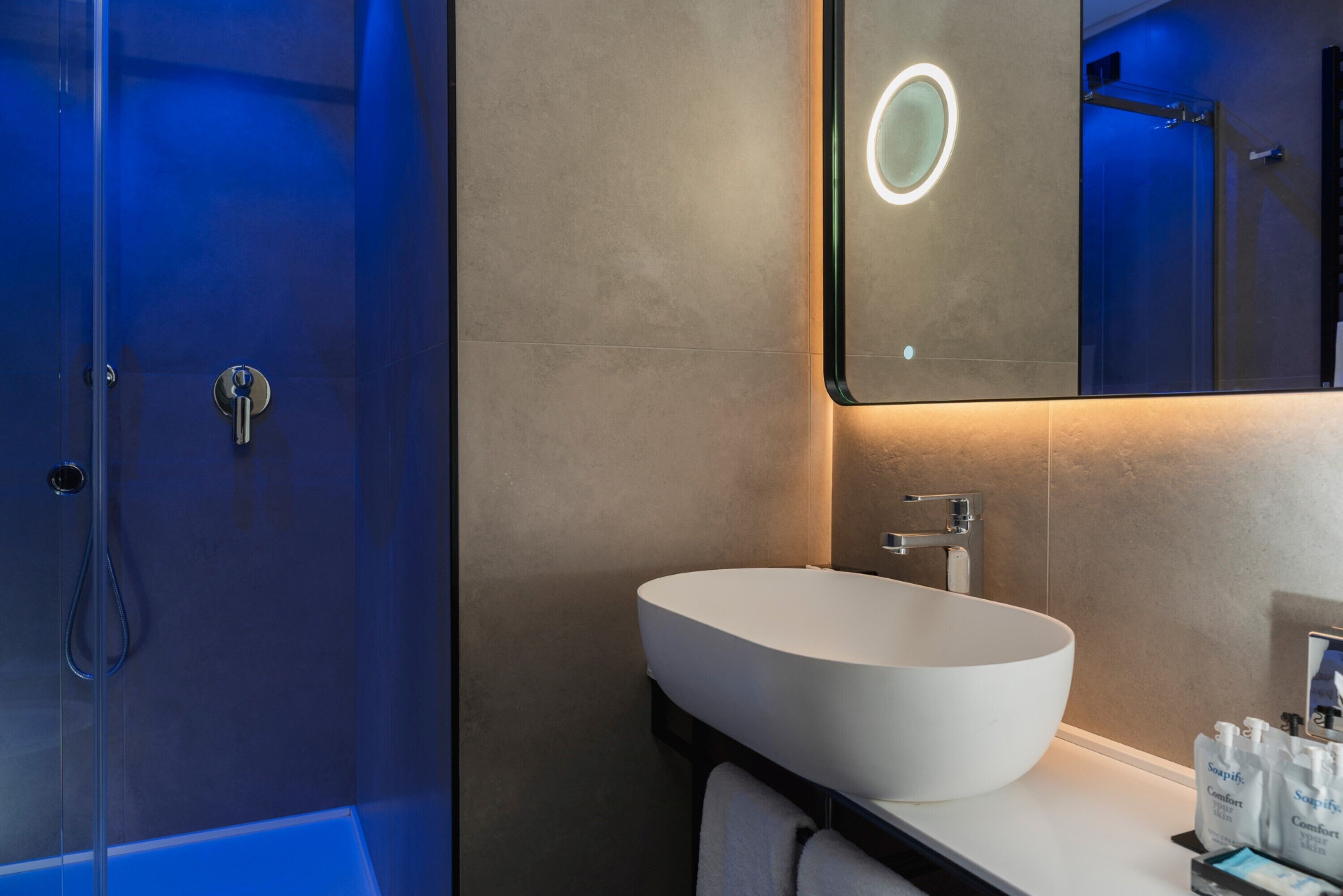 A modern bathroom with a large white sink, a mirror with a circular light, and a shower area.