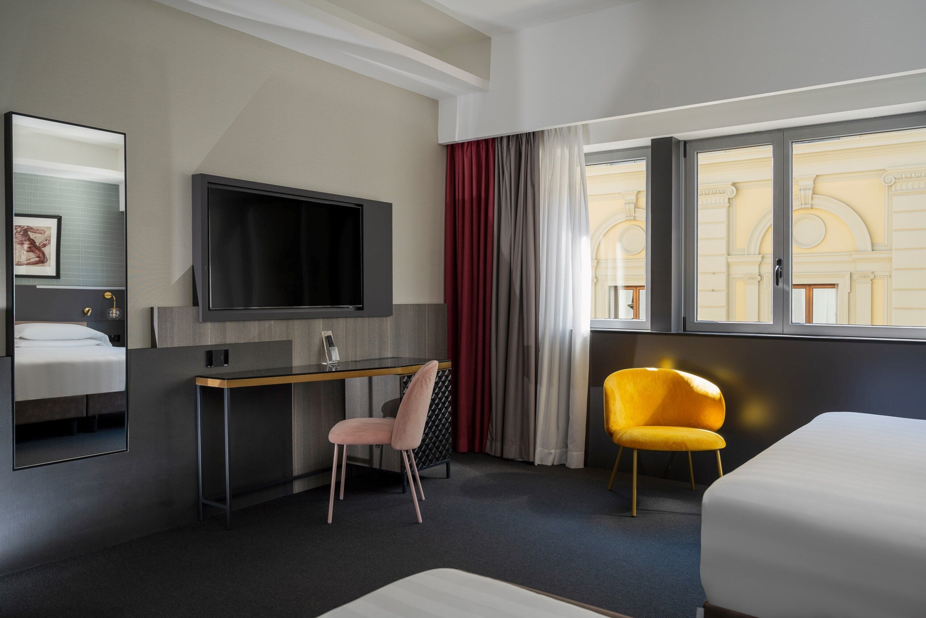 A hotel room with a bed, a desk with a chair, a TV, and a window with curtains.