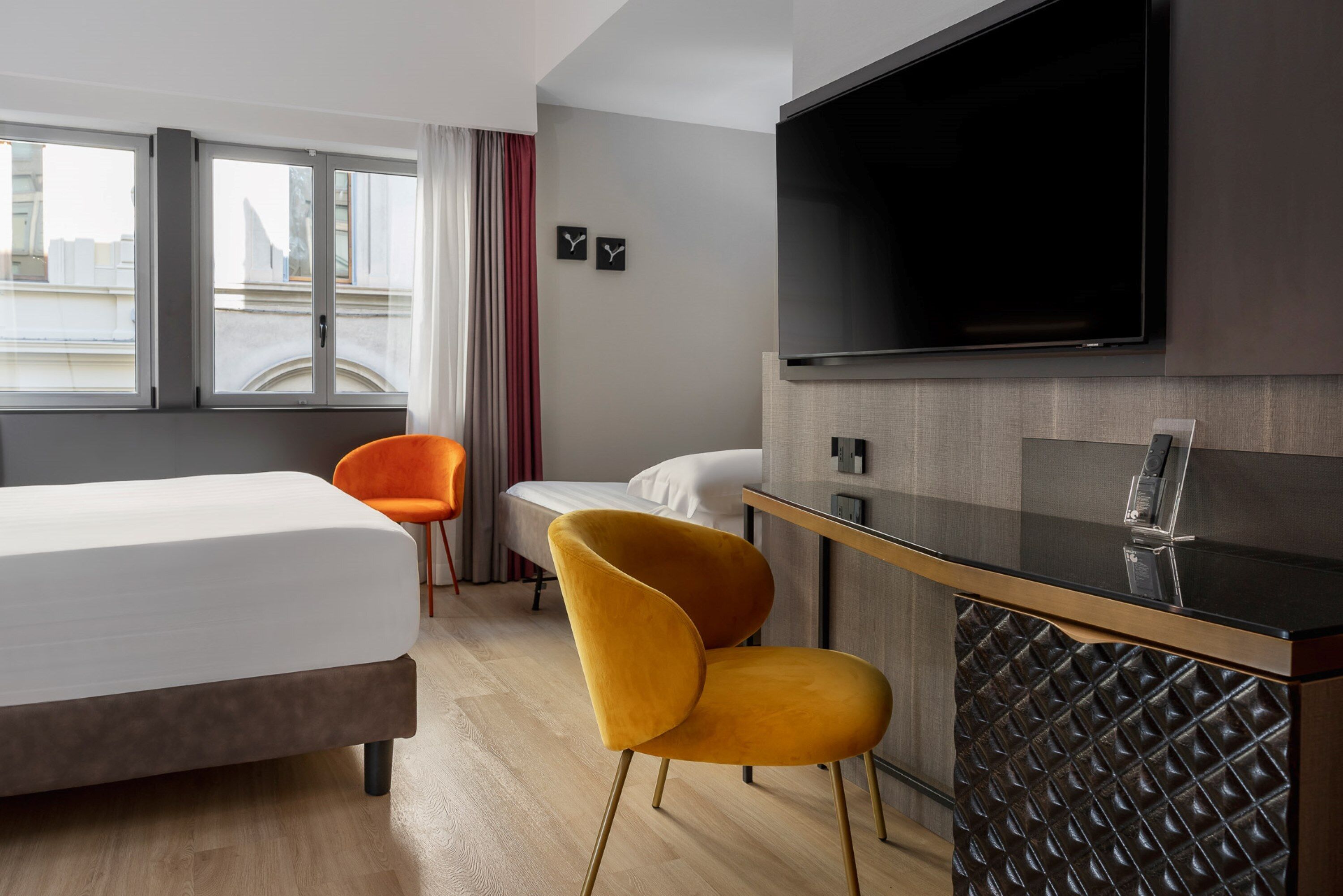 Ein modernes Hotelzimmer mit einem Bett, einem Schreibtisch mit Flachbildfernseher, einem Sessel und Blick auf die Stadt durch große Fenster.