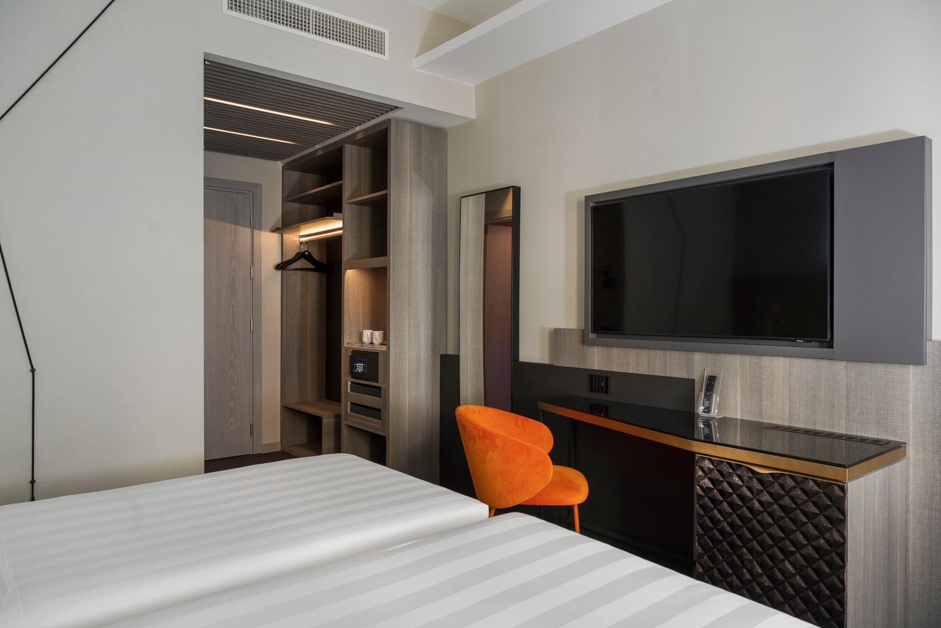 A modern hotel room with a bed, a desk with a chair, a TV mounted on the wall, and a walk-in closet.