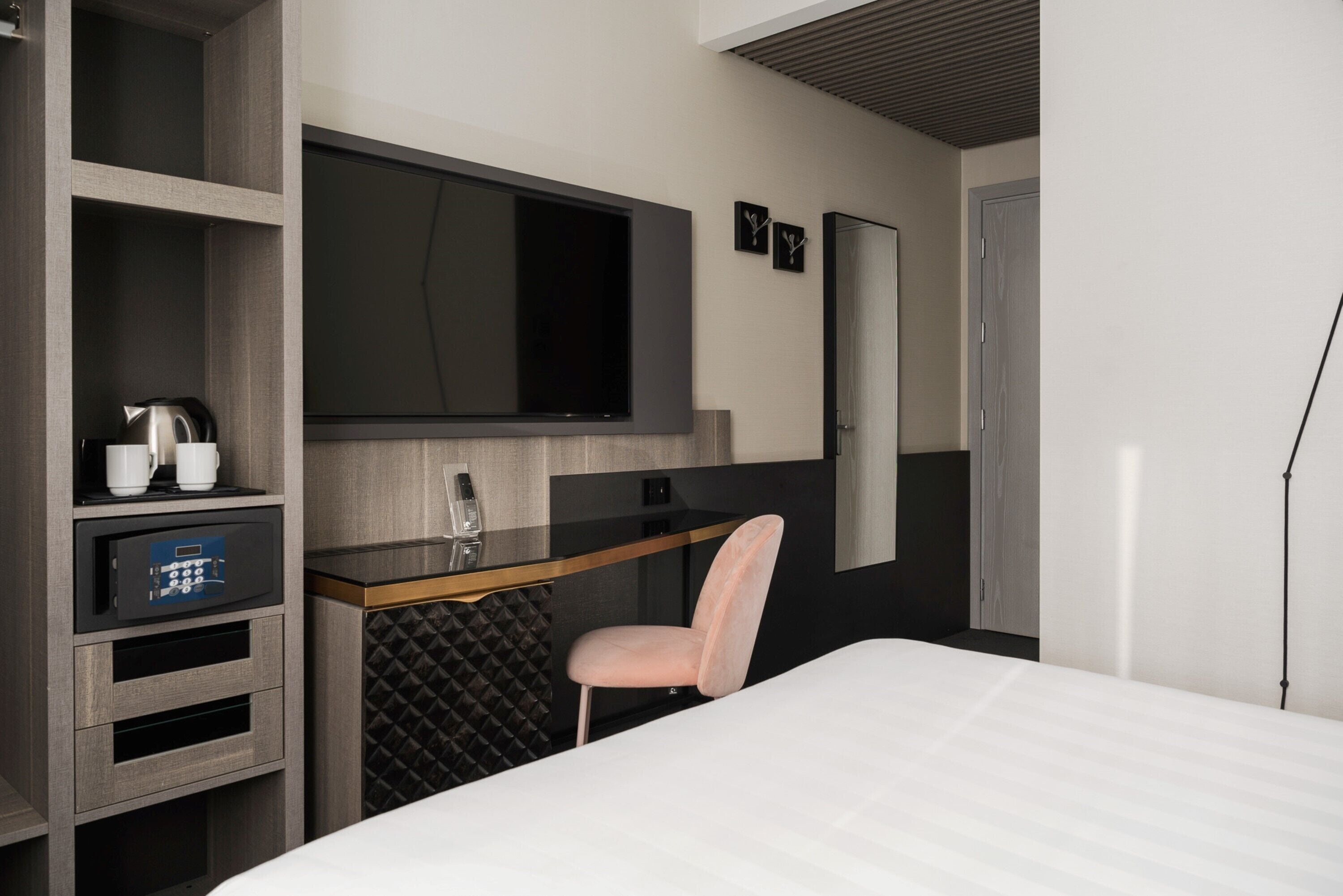 A modern hotel room with a bed, a desk with a chair, a television, a safe, and a wardrobe.
