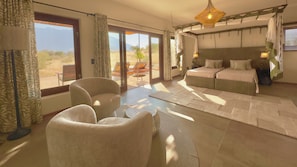Una habitación amplia con una cama grande, un sofá y una silla. Se aprecia vista al exterior a través de las ventanas.