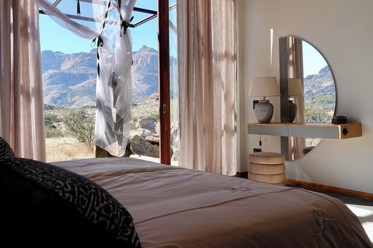 Un dormitorio con un ventanal grande, una cama con almohadas estampadas, una mesita de noche con lámpara y vistas a la montaña.
