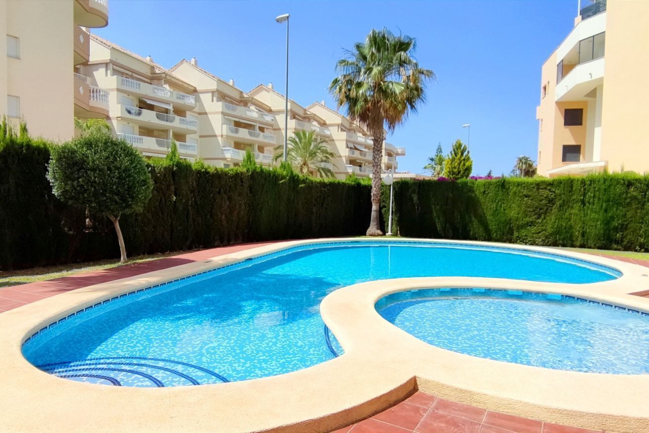 A swimming pool with a curved design, surrounded by a hedge and a building with balconies.