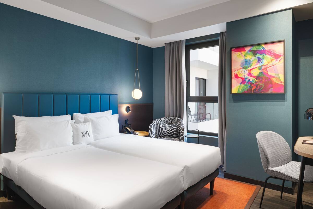 Ein Hotelzimmer mit einem großen Bett, einem Schreibtisch, einem Stuhl, einem Fenster mit Vorhängen und einem farbenfrohen abstrakten Gemälde an der Wand.