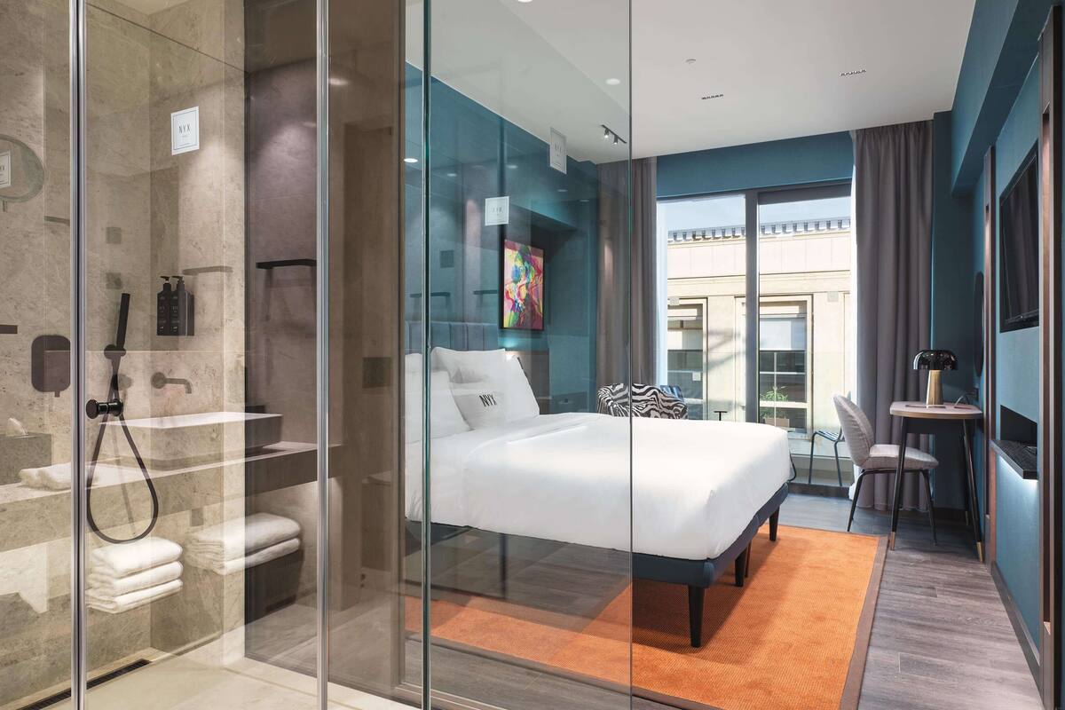 Ein modernes Hotelzimmer mit einer Glasabtrennung, die Duschbereich und Schlafbereich voneinander trennt. Das Zimmer verfügt über ein großes Bett, eine kleine Sitzecke mit Tisch und einen Ausblick nach außen durch das Glas.
