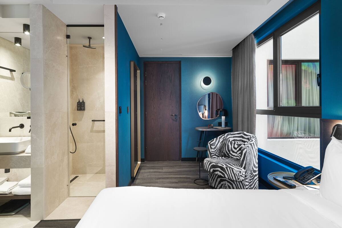 Ein modernes Hotelzimmer mit Dusche, einem Bett, einem zebra-gemusterten Sessel, einem runden Spiegel und Blick durch ein großes Fenster nach draußen.