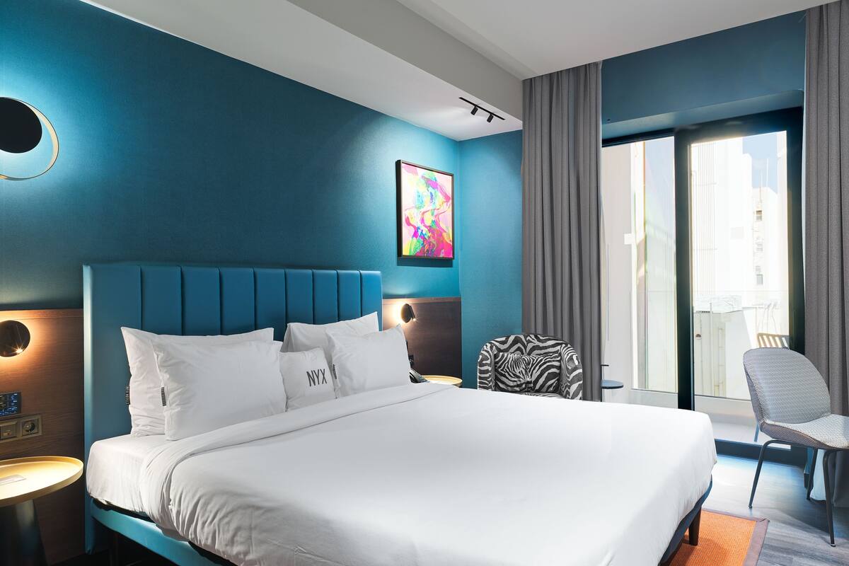 Ein modernes Hotelzimmer mit einem großen Bett, einer runden Wanduhr, einem zebra-muster Stuhl und Blick auf die Stadt durch das Fenster.
