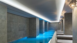 A modern indoor swimming pool with a sleek design, featuring a long, narrow pool area with a clear blue water surface, surrounded by stone walls and equipped with comfortable seating along the side.