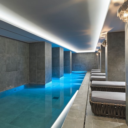A modern indoor swimming pool with a sleek design, featuring a long, narrow pool area with a clear blue water surface, surrounded by stone walls and equipped with comfortable seating along the side.