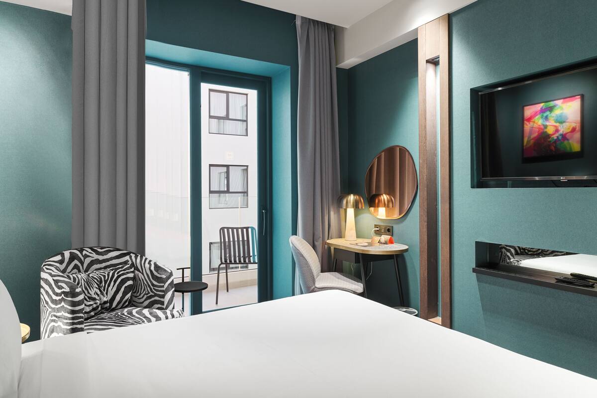 Ein Hotelzimmer mit Bett, einem zebraartig gemusterten Sessel, einem runden Tisch mit Lampe, einem an der Wand befestigten Fernseher und Blick ins Freie durch eine Tür.