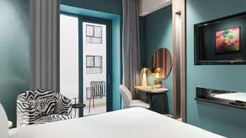 A hotel room with a bed, a zebra-patterned armchair, a round table with a lamp, a TV mounted on the wall, and a view of the outside through a door.