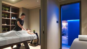 Sala de massagem com mesa de massagem, prateleira com plantas em vasos e corredor iluminado em azul que leva a uma área de spa.