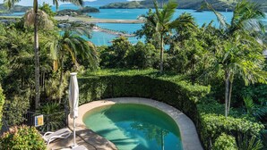 Piscina rodeada por vegetação exuberante e vista para uma área costeira com barcos e colinas.