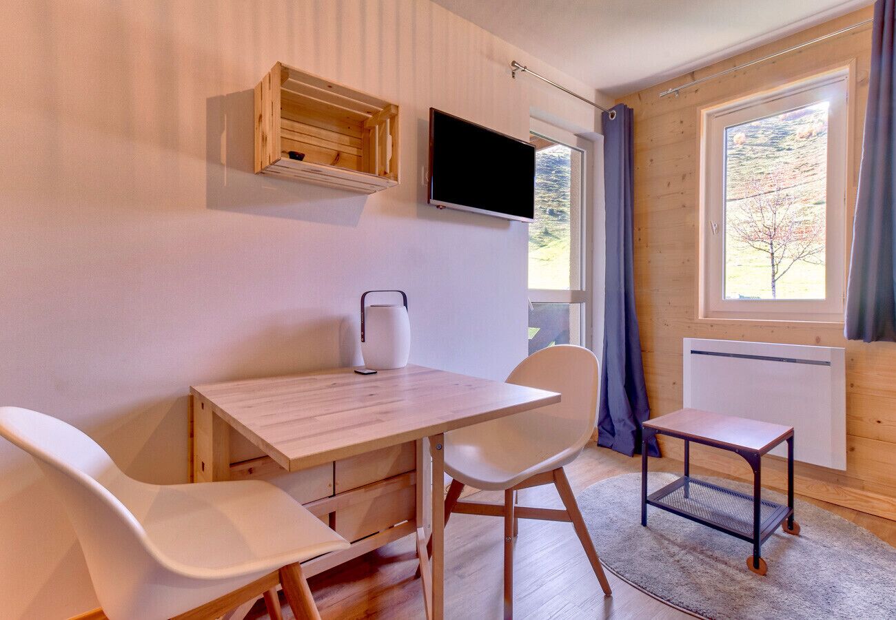 Una habitación con una mesa de madera y dos sillas blancas, una mesita auxiliar blanca, un televisor montado en la pared y una ventana con cortinas.
