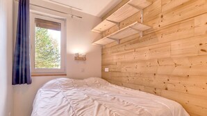 Ein Schlafzimmer mit Holz-Kopfteil, einem Bett mit weißer Decke, einem Fenster mit Blick auf Bäume und einem Vorhang links.