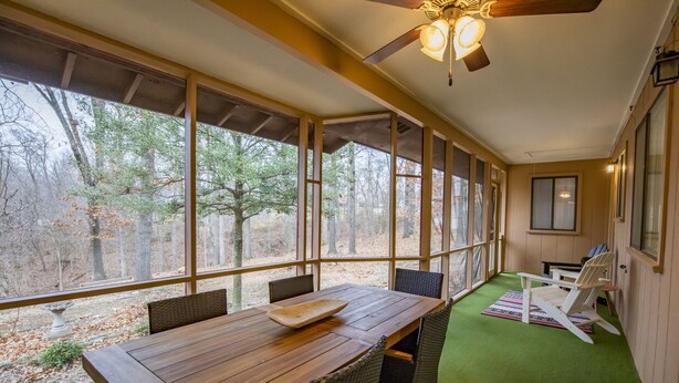 A covered patio with a wooden table and chairs, large windows, and a ceiling fan.