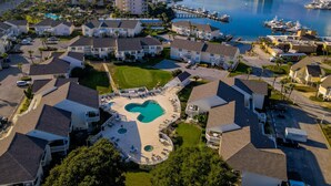 A residential area with a central swimming pool surrounded by houses, a marina with boats, and a beach in the background.