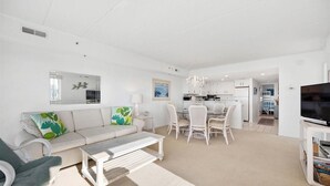 Een ruime woonkamer met een bank, salontafel en een eethoek met stoelen. Er is een televisie en een boekenkast in de kamer.