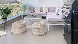 A balcony with a sofa, two woven ottomans, a grill, and a view of a cityscape. - ALL COMFORT, SUPERB VIEW, large TERRACE Los Flamingos Ideal families (Estepona)