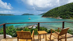 Une terrasse en bois avec deux chaises et une table, d’où l’on peut admirer une mer turquoise et un voilier, le tout entouré d’une végétation luxuriante et de collines.