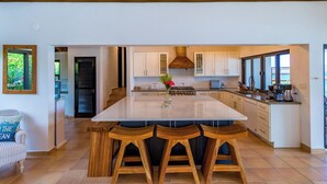 Une cuisine moderne dotée d’un grand îlot central, de tabourets en bois, de meubles blancs et offrant une vue sur l’extérieur.