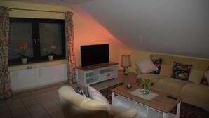Ein Wohnzimmer mit einer Couch, einem Fernseher, einem Couchtisch und einem Fenster mit Vorhängen.