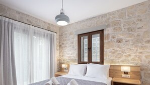 Une chambre avec un mur en pierre, un grand lit recouvert d’une courtepointe, deux tables de chevet avec des lampes et une fenêtre avec des rideaux.