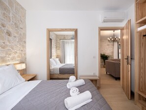 Une chambre d’hôtel avec un mur en pierre, un lit recouvert d’une courtepointe grise matelassée, une tête de lit en bois, une table de chevet avec une lampe, un miroir, une armoire en bois et une porte donnant sur une salle de bain.