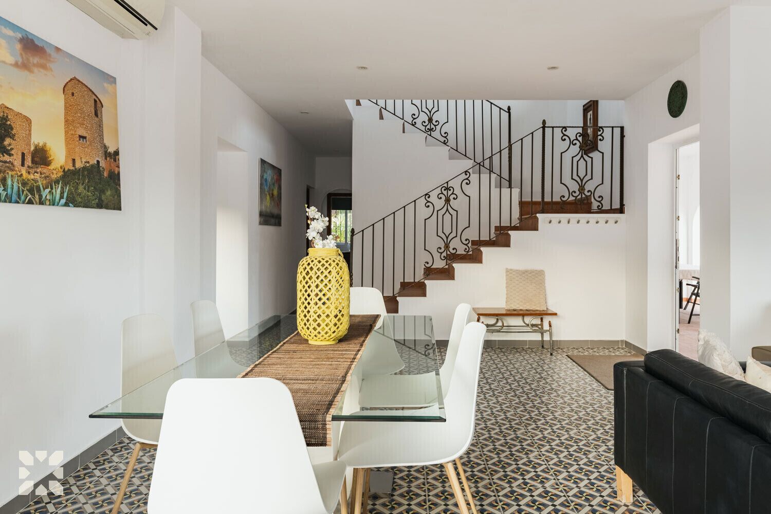 A modern living room with a glass dining table, white chairs, a black sofa, a staircase with ornate railings, and a painting of a building.