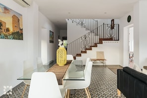 A modern living room with a glass dining table, white chairs, a black sofa, a staircase with ornate railings, and a painting of a building.